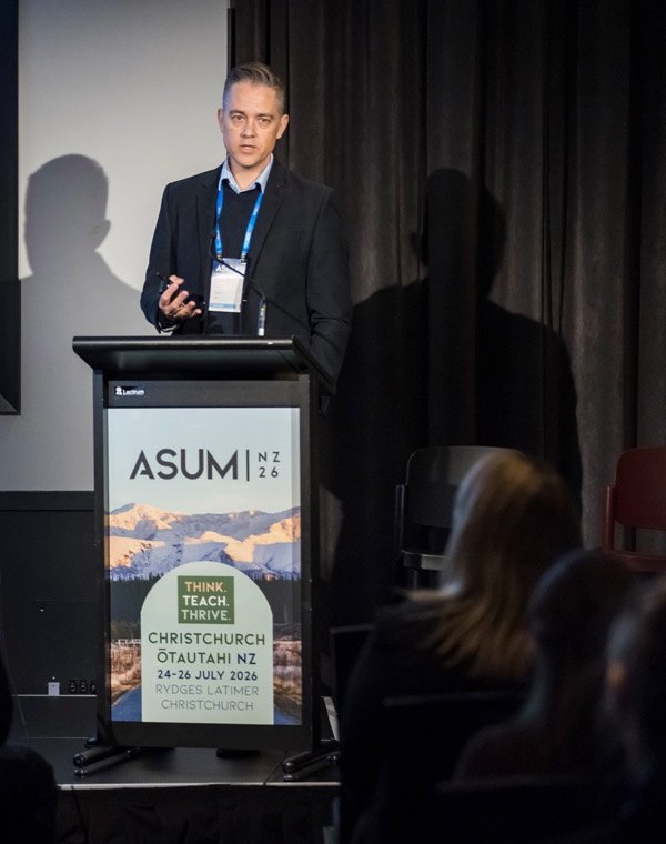 Tristan Reddan presenting at a lectern bearing the ASUM NZ 2026 conference banner, Christchurch.