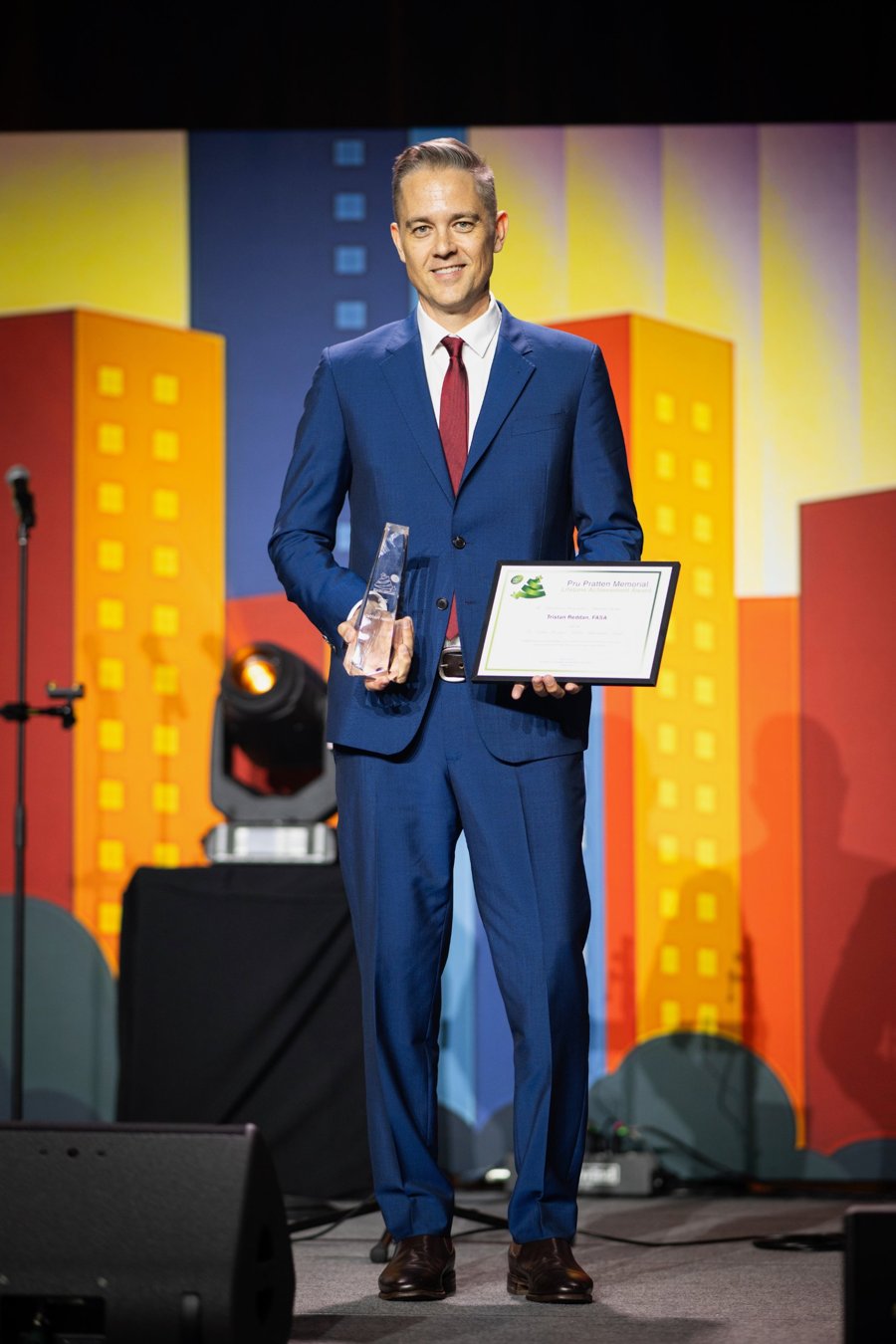 Tristan on stage holding the Pru Pratten Memorial Lifetime Achievement Award trophy and certificate.