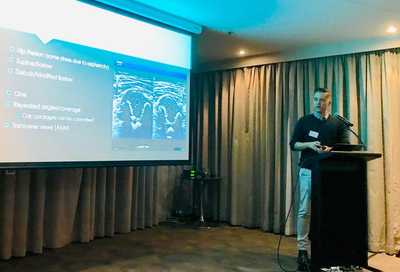 Tristan presenting hip ultrasound technique slides at a lectern in a function room.