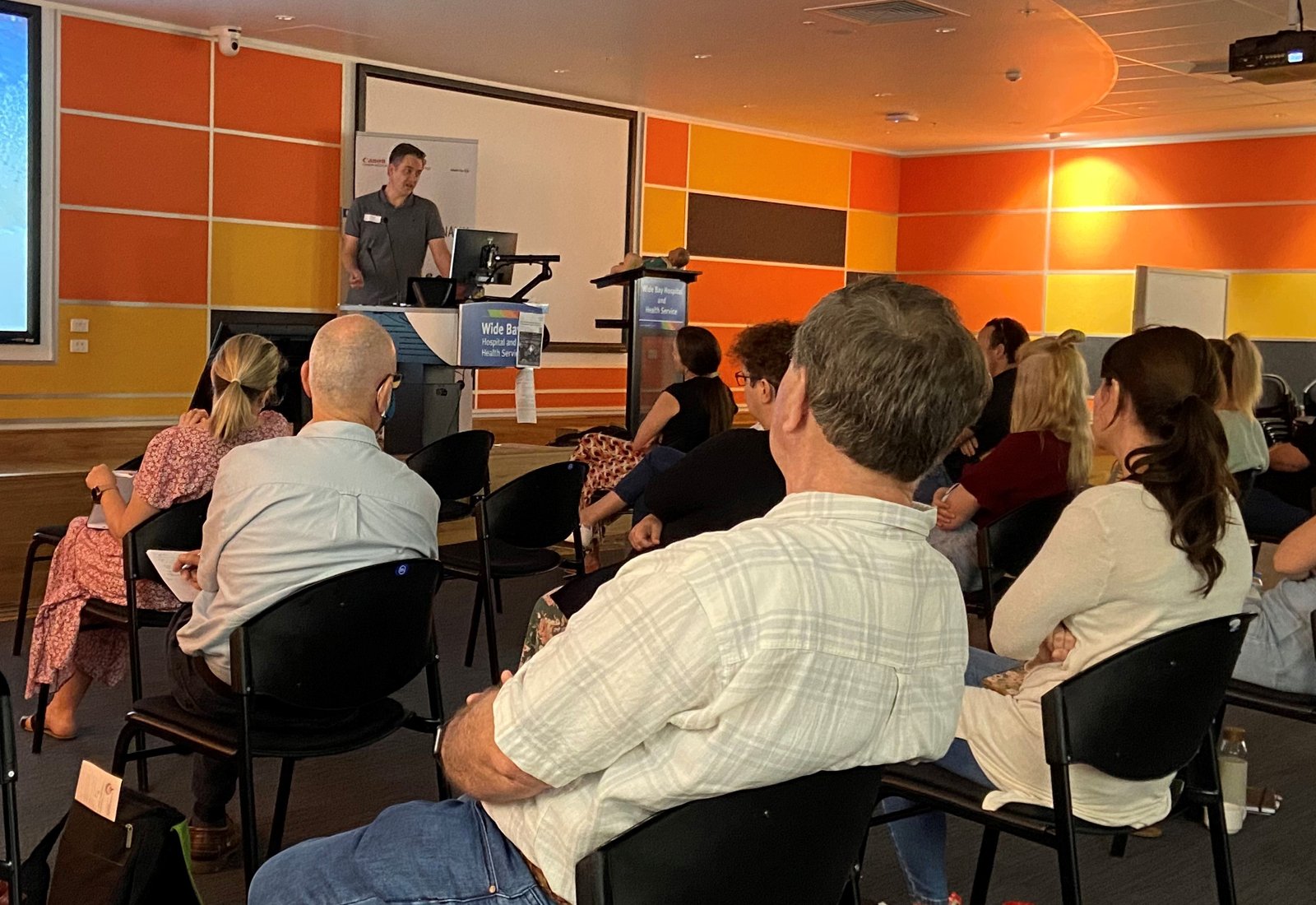 Tristan presenting at a lectern in a regional hospital education room, with the audience seated in the foreground.