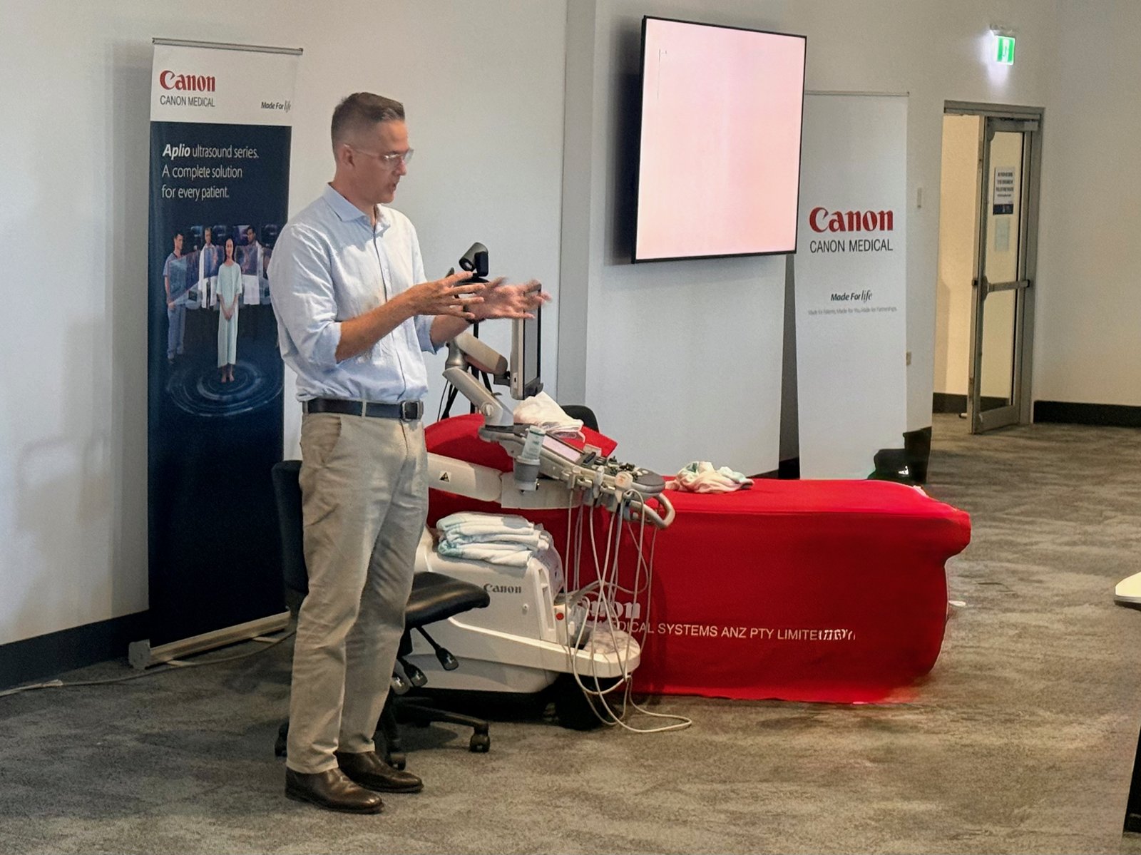 Tristan presenting in front of Canon Medical banners with an ultrasound machine and examination couch beside him.